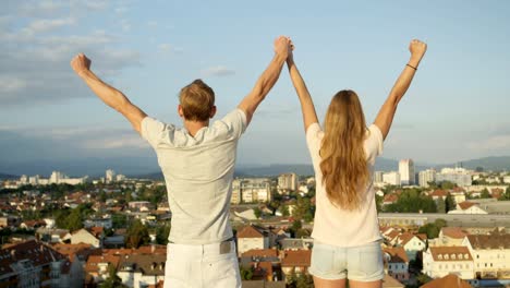 CLOSE-UP:-Young-couple-standing-on-rooftop-above-the-city,-raising-hands-in-sky