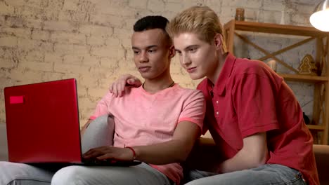 Two-multinational-homosexual-friends-sit-on-the-couch,-use-a-laptop,-watch-a-funny-video-on-the-Internet,-laughing.-Home-cosiness,-family,-internet-concept.-60-fps