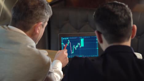 Two-colleagues-a-mature-man-and-young-man-discuss-trading-with-cryptocurrency-on-laptop-and-drink-coffee