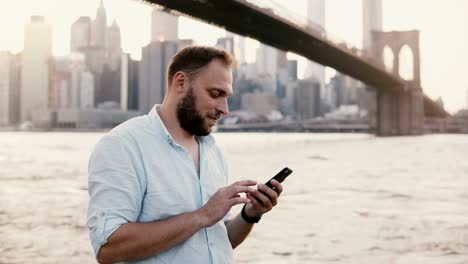 Glücklich-kaukasischen-freier-Unternehmer-Eingabe-Nachricht-auf-Smartphone,-lächelnd-in-der-Nähe-von-Brooklyn-Bridge-New-York-City-4K