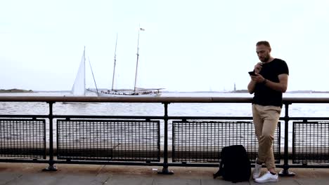 Young-handsome-man-standing-on-the-pier-near-the-fence-and-using-smartphone-near-the-water,-browsing-the-Internet