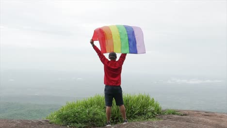 hombre-levantar-bandera-LGBTI-de-color-arco-iris-ondeando-en-duro-viento-en-el-mirador-de-alta-montaña