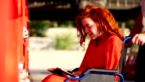 Young-Man-Rolls-His-Girlfriend-In-A-Wheelchair-To-The-Bookshelf,-She-Takes-Her-Favorite-Book-And-Tells-Him-About-This-Book,-Outdoors