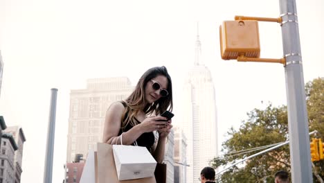 Trabajador-independiente-femenino-europeo-feliz-con-bolsas-de-la-compra-de-gafas-de-sol-sonriente,-usando-la-aplicación-para-smartphone-en-Nueva-York