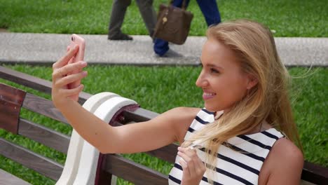 Junge-schöne-schlanke-Frau-mit-langen-blonden-Haaren-in-schwarzen-und-weißen-Kleid-machen-Selfie-auf-Handy-über-Hintergrund-des-Parks.