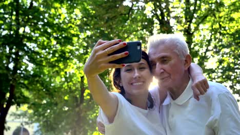 Una-niñera,-enfermera,-cuidar-de-los-ancianos,-una-chica-(mujer)-y-el-abuelo-se-sientan-en-un-libro-de-bolsillo-y-hacer-selfie,-en-el-parque.