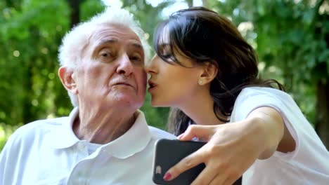 Una-niñera,-enfermera,-cuidar-de-los-ancianos,-una-chica-(mujer)-y-el-abuelo-se-sientan-en-un-libro-de-bolsillo-y-hacer-selfie,-en-el-parque.