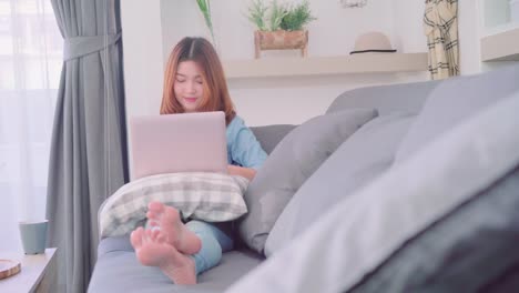 Portrait-of-beautiful-attractive-young-smiling-Asian-woman-using-computer-or-laptop-while-lying-on-the-sofa-when-relax-in-living-room-at-home.-Enjoying-time-lifestyle-women-at-home-concept.