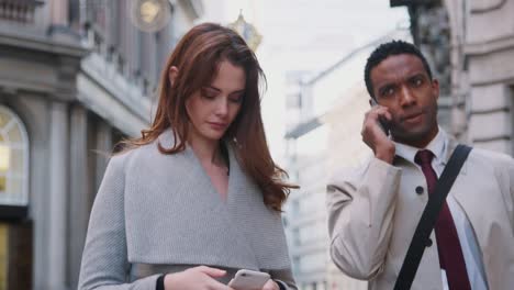 Millennial-white-woman-standing-on-the-street-using-smartphone,-young-black-businessman-talking-on-phone-walking-past,-close-up,-low-angle