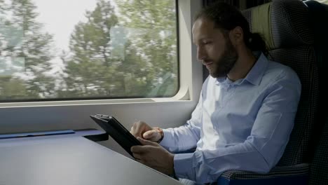 Focused-young-entrepreneur-swiping-and-scrolling-on-tablet-touchscreen-surfing-on-internet-next-to-window-in-moving-train