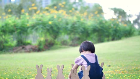 Niedliches-kleines-Kindermädchen-am-Ostersamstag.-Mädchen-jagt-nach-Ostereiern-auf-dem-Rasen-und-Hase-Made-of-paper-in-der-Natur-oder-Park-und-Sonnenlicht.-Zeitlupe