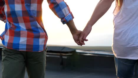 Vista-trasera-de-la-pareja-masculina-sosteniendo-las-manos-entre-sí-y-jogging-en-la-terraza-de-la-azotea.-Jóvenes-chicos-gays-corriendo-al-borde-de-la-azotea-y-levantando-los-brazos.-Hermoso-paisaje-urbano-en-el-fondo.-Cámara-lenta