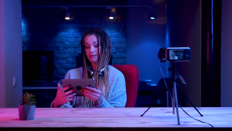 Closeup-shoot-of-young-female-blogger-with-dreadlocks-in-headphones-streaming-live-using-the-tablet-and-showing-green-chroma-screen-to-the-camera-with-the-neon-background