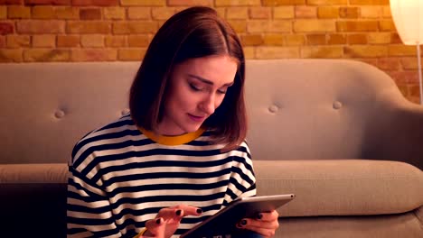 Closeup-portrait-of-young-pretty-girl-typing-on-the-tablet-and-smiling-happily-sitting-on-the-floor-in-a-cozy-apartment-indoors