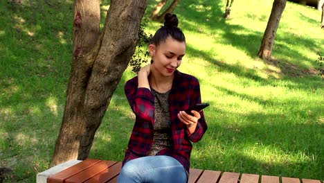 Young-beautiful-girl-in-a-plaid-shirt-sits-on-a-bench-in-the-park,-actively-rewritten-in-internet-chat