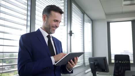 Cheerful-businessman-using-tablet-and-leaning-against-office-windows