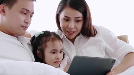 Asian-family-sitting-on-bed-laughing-and-looking-in-digital-tablet-while-lying-in-bedroom