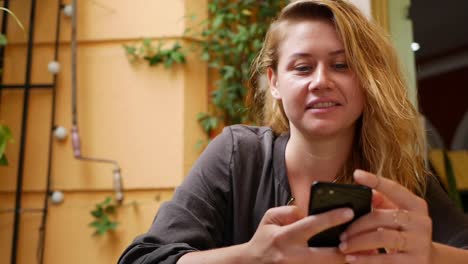 Happy-girl-uses-a-smartphone,-writes-a-message,-chatting.-Young-woman-looking-for-information-in-the-phone.-Sitting-in-restaurant-with-friends.-Breakfast-time