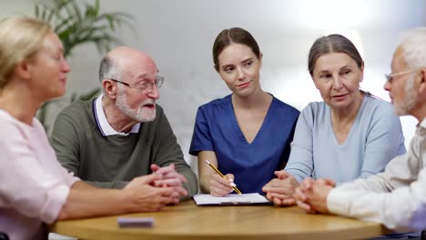 Gruppe-von-vier-Senioren,-die-im-Pflegeheim-am-Tisch-sitzen-und-reden.-Junge-Krankenschwester-in-Uniform-oder-Therapeutin,-die-Notizen-in-medizinischen-Dokumenten-auf-der-Zwischenablage-macht