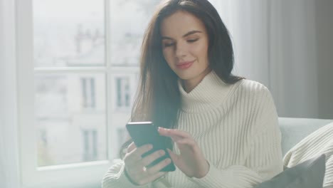 Schöne-junge-Frau-mit-Smartphone-glücklich,-während-sitzen-auf-Stuhl.-Sinnliche-Mädchen-tragen-gestrickte-Pullover,-Surfs-Internet,-Beiträge-auf-Social-Media,-Teilen-Bild-beim-Entspannen-in-gemütlichen-Wohnung