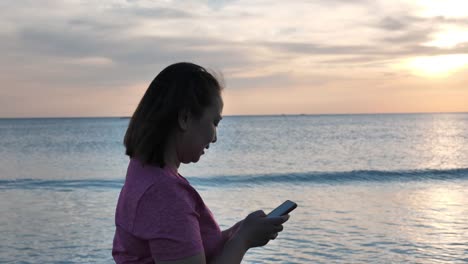 Nahaufnahme-Seitenblick-asiatische-Frauen-entspannen-zu-Fuß-am-Strand-und-verwenden-ein-Handy-am-Abend,-Sonnenuntergang.-Urlaubs--und-Reisekonzept.-Zeitlupe