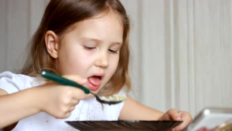 Kind-isst-Suppe-mit-einem-Löffel-und-schaut-Video-auf-einem-Smartphone.-Closeup-Baby-Mädchen-sitzt-am-Tisch-der-Hausmannskost-und-Essen-essen