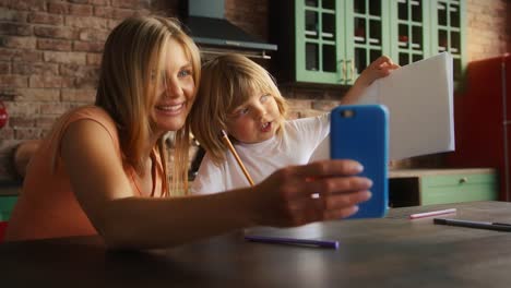 Lächelnde-Mutter-und-Kind-genießen-Online-Videoanruf-auf-dem-Smartphone,-während-Hausaufgaben-in-der-Schule-Copybook.-Sie-sitzen-in-der-Küche-am-Tisch.-Nahaufnahme