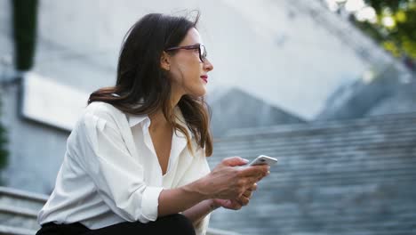 Female-entrepreneur-in-glasses,-formal-attire.-Sitting-on-stairs-and-typing-on-smartphone,-working-online-outdoors.-Business-concept.-Close-up