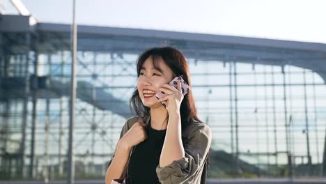 Muy-alegre-joven-mujer-asiática-con-el-pelo-oscuro-hablando-por-teléfono-y-sonrisa-sinceramente-cerca-del-edificio-del-aeropuerto-en-el-día