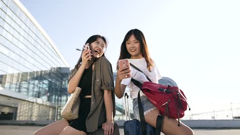 Happy-good-looking-25-years-old-asian-girlfriends-sitting-on-their-suitcases-and-using-smartphones-near-the-modern-airport-terminal