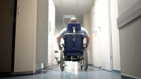 Rear-view-of-disabled-man-pushes-himself-in-wheelchair-down-hospital-corridor