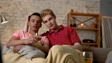 Multinational-gay-couple-sitting-on-couch-covered-with-a-warm-blanket,-watch-TV,-use-the-remote-control,-look-at-the-camera.-Homeliness,-romantic-evening,-background,-hugs,-happy-LGBT-family-concept.-60-fps