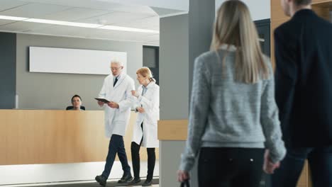 In-the-Hospital,-Busy-Doctors-Talk,-Using-Tablet-Computer-While-Walking-Through-the-Building.-In-the-Background-Patient-Talks-with-Receptionist.-New-Modern-Fully-Functional-Medical-Facility.