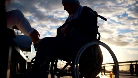 Silhouette-of-young-woman-with-disabled-man-in-a-wheelchair-talking-at-sunset-outdoors