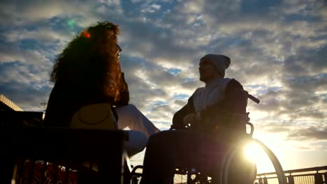 Young-woman-with-disabled-man-in-a-wheelchair-talking-outdoors-at-sunset