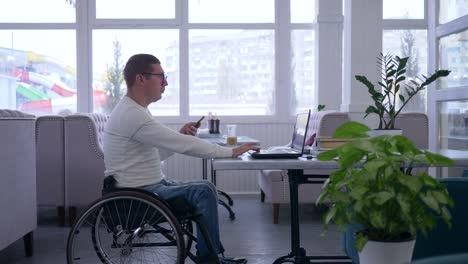 remote-business-management,-handicapped-man-in-wheelchair-uses-a-cellphone-sitting-at-table-with-laptop-in-cafe-on-the-background-of-large-window