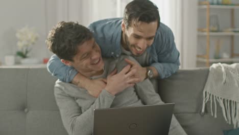 Süße-männliche-Queer-paar-verbringen-Zeit-zu-Hause.-Junger-Mann-verwendet-einen-Laptop,-seine-Partnerin-von-hinten-kommt-und-sanft-umarmt-ihn.-Sie-lachen-und-Hände-berühren.-Zimmer-ist-Modern-eingerichtet.