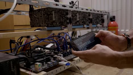 Trader-man-holding-his-smartphone-near-the-gpu-cryptocurrency-mining-rig-with-crypto-mining-software-program-running-on-the-digital-display-mobile-phone