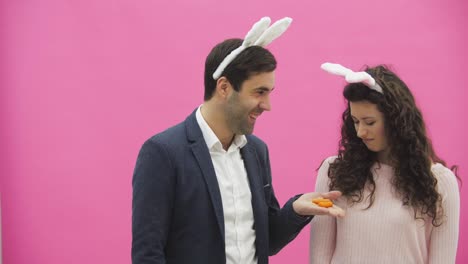 Young-couple-are-beautiful-on-pink-background.-During-this-time,-they-are-dressed-in-rabble-ears.-Looking-at-each-other.-A-man-shows-carrots,-his-wife-does-not-like-the-size-of-carrots-and-she-throws-out.