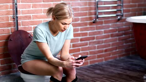 Mujer-joven-confiada-sentada-en-el-inodoro-en-el-baño-usando-un-teléfono-inteligente-de-tiro-medio-largo