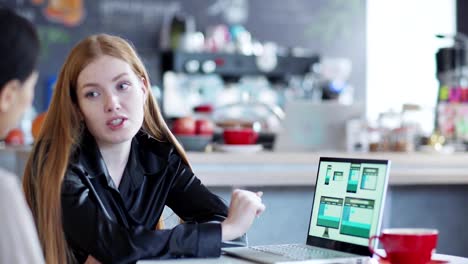 Zoom-out-of-two-young-businesswomen-discussing-website-templates-for-different-devices-presented-on-laptop-screen-sitting-at-table-in-cafe,-focus-on-woman-explaining-design-to-Asian-colleague