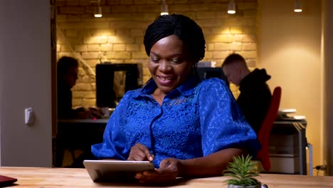 Closeup-shoot-of-happy-adult-african-american-businesswoman-using-the-tablet-and-laughing-cheerfully-in-the-office-indoors