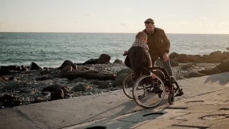 Hombre-amoroso-está-girando-silla-de-ruedas-con-la-mujer-joven-en-la-costa-del-mar,-regocijo