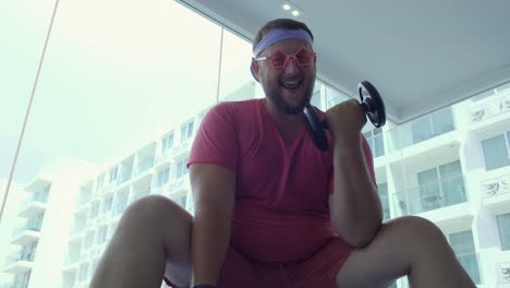 Hombre-gordo-divertido-en-gafas-rosas-y-en-una-camiseta-rosa-se-dedica-con-mancuernas-en-una-bola-de-ajuste-en-el-gimnasio-que-representa-a-una-chica