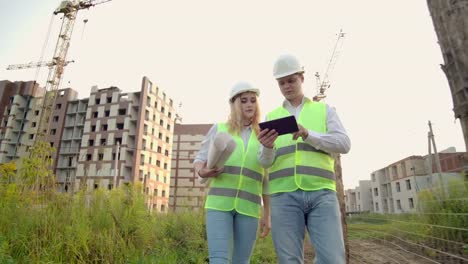 Negocios,-construcción,-industria,-tecnología-y-concepto-de-personas---constructor-sonriente-en-hardhat-con-tableta-PC-ordenador-junto-con-la-mujer-con-dibujos-de-constructores-en-el-sitio-de-construcción.