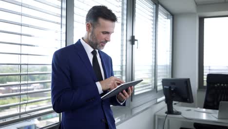 Smiling-businessman-using-tablet-and-leaning-against-office-window