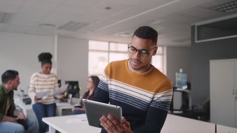 Joven-empresario-africano-creativo-ocupado-en-el-uso-de-la-tableta-digital-mientras-sus-colegas-del-equipo-discuten-el-trabajo-en-segundo-plano