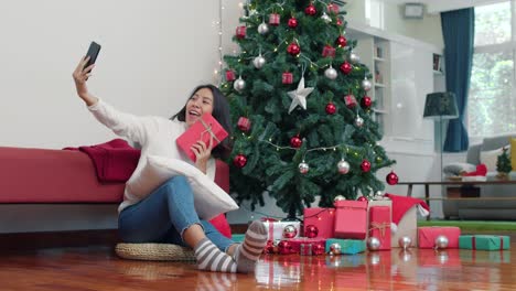 Asiatische-Frauen-feiern-Weihnachtsfest.-Weibliche-teen-entspannen-glücklich-halten-Geschenk-und-mit-Smartphone-Selfie-mit-Weihnachtsbaum-genießen-Xmas-Winterurlaub-im-Wohnzimmer-zu-Hause.