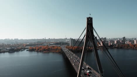 Busy-car-traffic-on-bridge-in-big-metropolis.-High-rise-buildings-and-plant-background.-Busy-city-promenade-in-city-center.-Concrete-bridge-with-steel-rope-tows.-Aerial-drone-view.-Kyiv,-Ukraine