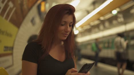 Atractiva-chica-pelirroja-con-pecas,-piercings-y-pelo-rojo-charlando-en-el-teléfono-inteligente-en-la-estación-de-metro,-durante-el-verano-soleado-en-París.-Fondo-subterráneo-borroso.-4K-UHD.-Viene-el-tren.